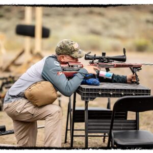 Man aiming a rifle equipped with a Rimfire Drop-In scope at an outdoor shooting range, supported by a shooting rest on a table, wearing camouflage cap and tactical gear.