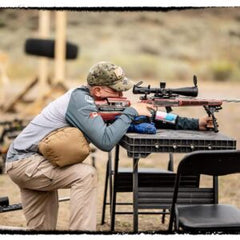 Man aiming a rifle equipped with a Rimfire Drop-In scope at an outdoor shooting range, supported by a shooting rest on a table, wearing camouflage cap and tactical gear.