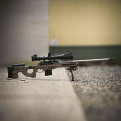 Precision rifle built with "Deadline 2.0 Action" featuring a black stock, mounted scope, and bipod on a smooth concrete and gravel surface.