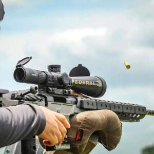 Close-up of a shooter aiming a rifle equipped with a scope and suppressor labeled Federal, featuring the custom barreled action Deadline 2.0 Action in an outdoor shooting range setting.