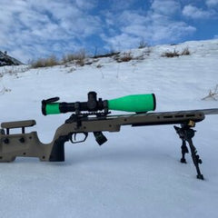 Rimfire Drop-In precision rifle with a green scope cover mounted on a tan custom barreled action, resting on a snow-covered landscape with a bipod for stability.